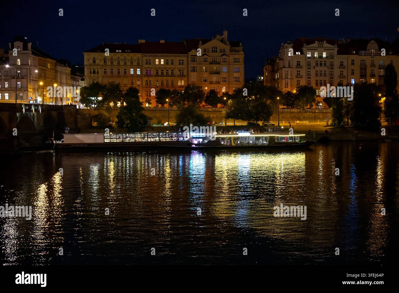 Vista notturna sul fiume Vltava con una nave da crociera illuminata e edifici storici lungo l'argine. Foto Stock