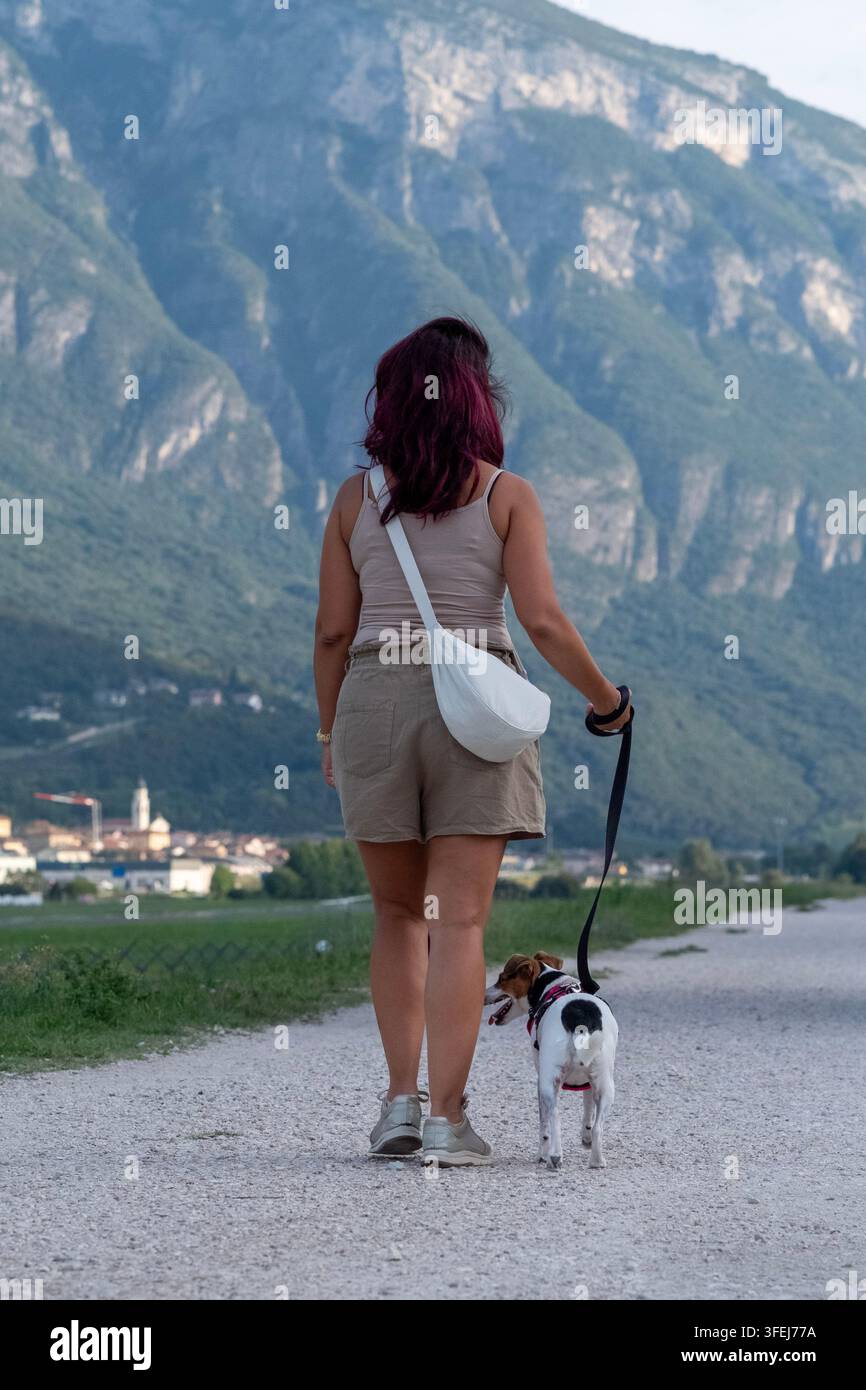 Una donna e il suo cane Jack Russell al guinzaglio, inquadrati da dietro, camminano lungo una strada di campagna, sullo sfondo il profilo di una montagna Foto Stock