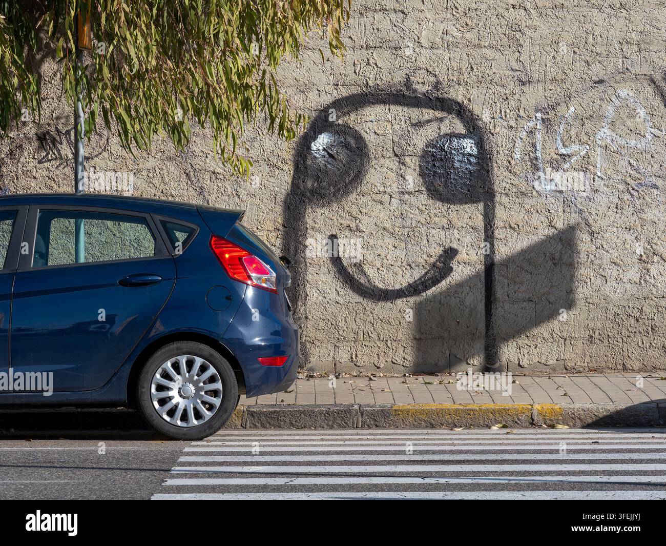 Ombra di un cartello stradale che crea un graffiti a forma di faccina sorridente su un muro urbano accanto a un'auto blu parcheggiata Foto Stock