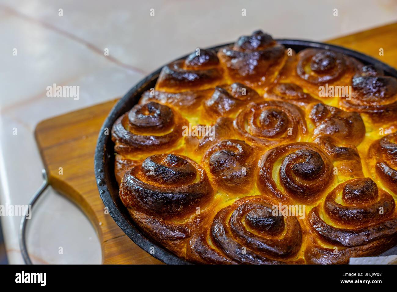 Rotoli di pasticceria appena sfornati con una crosta dorata sono disposti in una teglia nera circolare. La superficie lucida indica una glassa dolce, su uno sfondo di cucina sfocato. Foto Stock