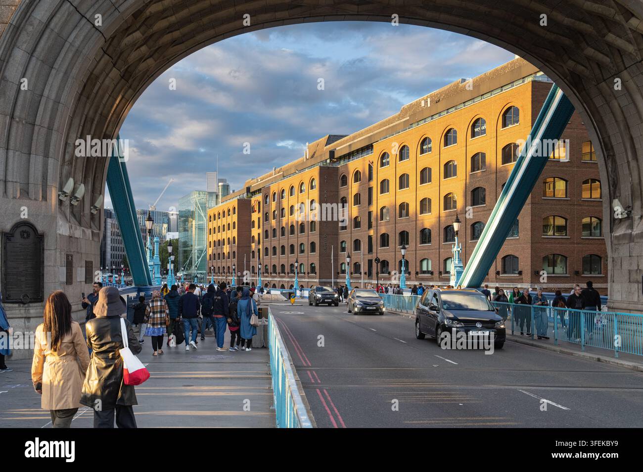 Pedoni e automobilisti attraversano il Tower Bridge di Londra, incorniciati dal suo arco di pietra, passando davanti all'edificio illuminato dal sole degli St Katharine Docks. Foto Stock