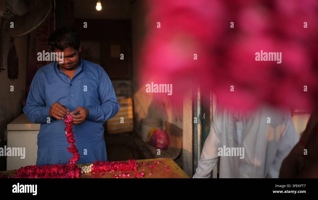 Shahdara, Punjab, Pakistan - 28 ottobre 2026: Vista di un uomo in camicia blu che crea una ghirlanda di petali di rosa dentro un negozio, con fiori rosa vivaci e Foto Stock