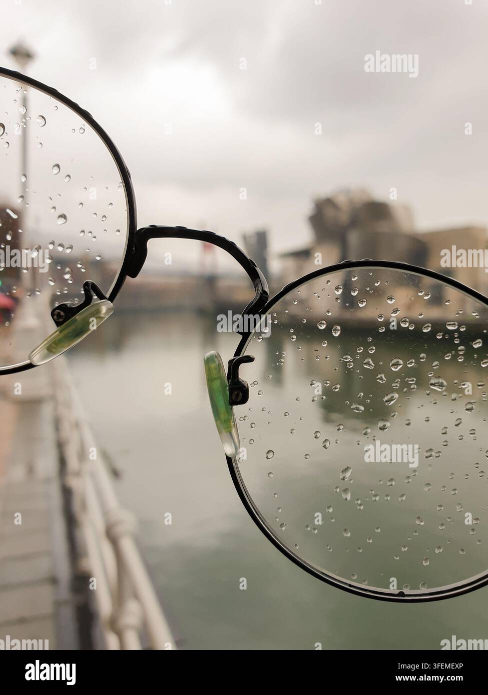 Spagna, Bilbao vista attraverso occhiali bagnati con lenti miopiche l'architettura sfocata del museo e il fiume con le ringhiere della passerella pedonale sotto una cielo nuvoloso Foto Stock