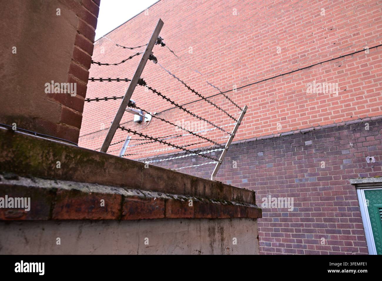 Muro di mattoni con barriera di sicurezza in filo spinato, porta metallica verde, telecamera di sorveglianza montata Foto Stock