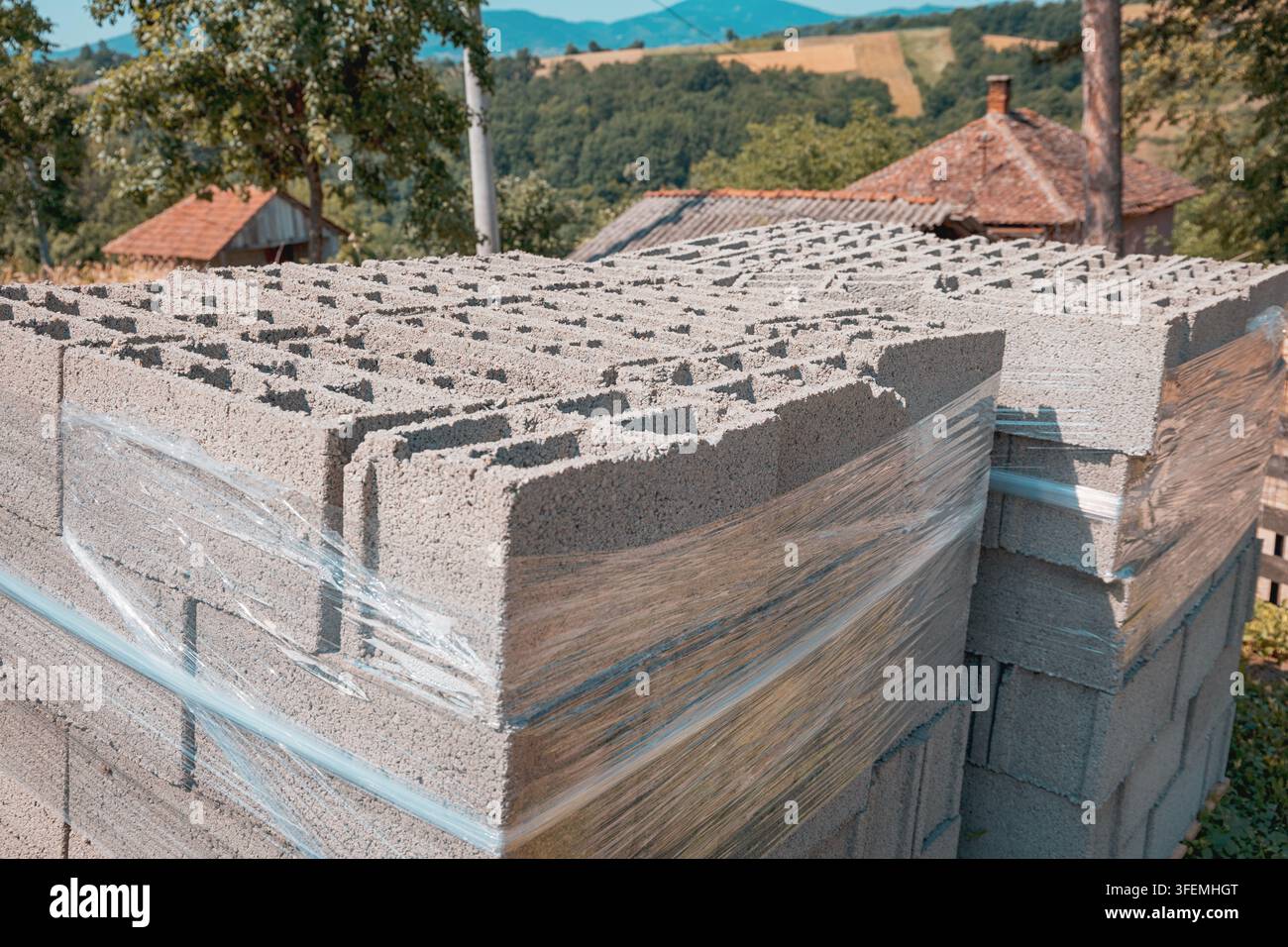 Pallette di blocchi di calcestruzzo cavi avvolti in fogli di plastica impilati all'aperto nella calda luce estiva, pronti per un progetto di costruzione rurale. Foto Stock