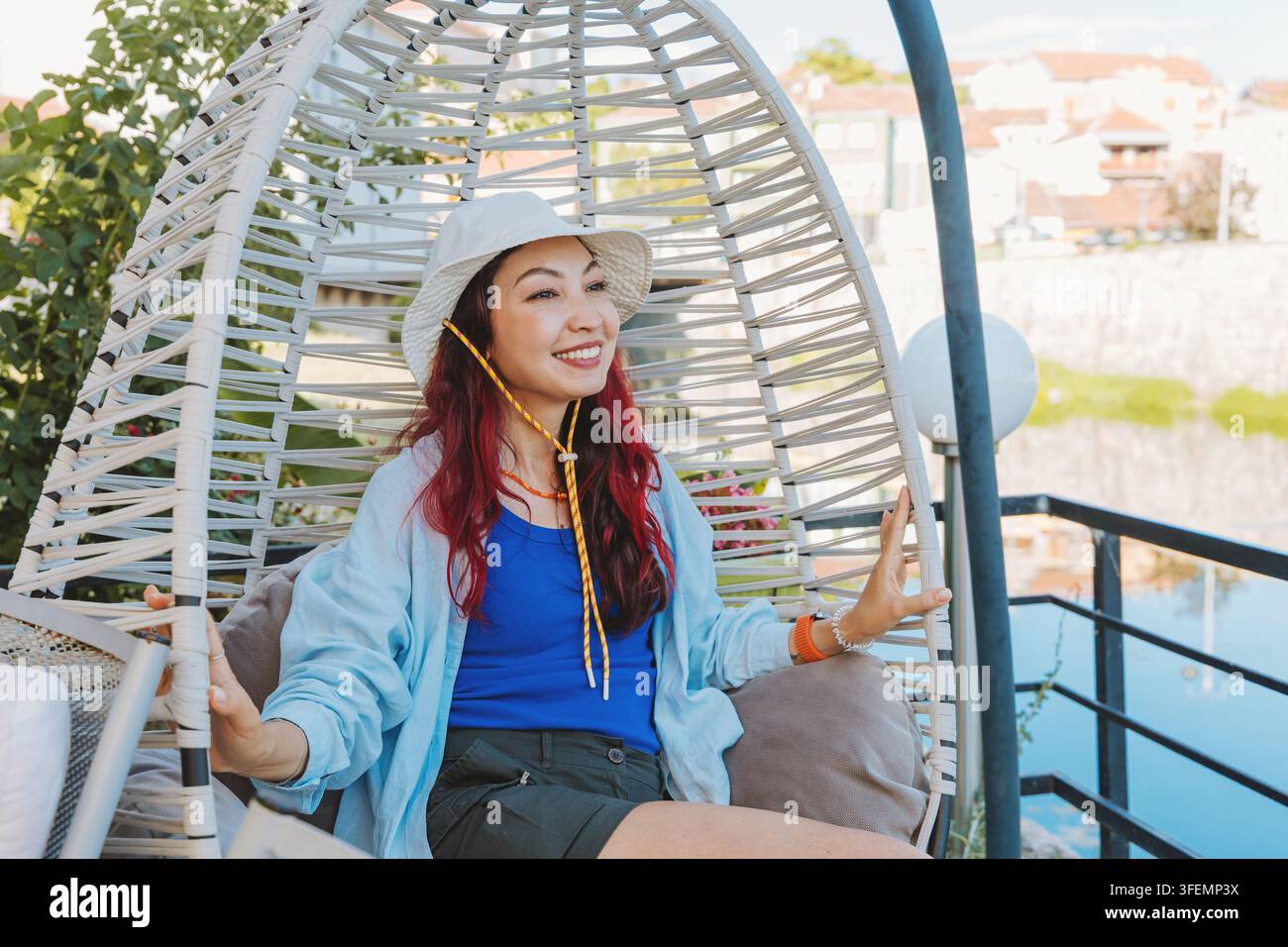 Donna felice con cappello da sole e abiti casual che si rilassa in una sedia a uovo oscillante all'aperto su una terrazza durante l'estate Foto Stock