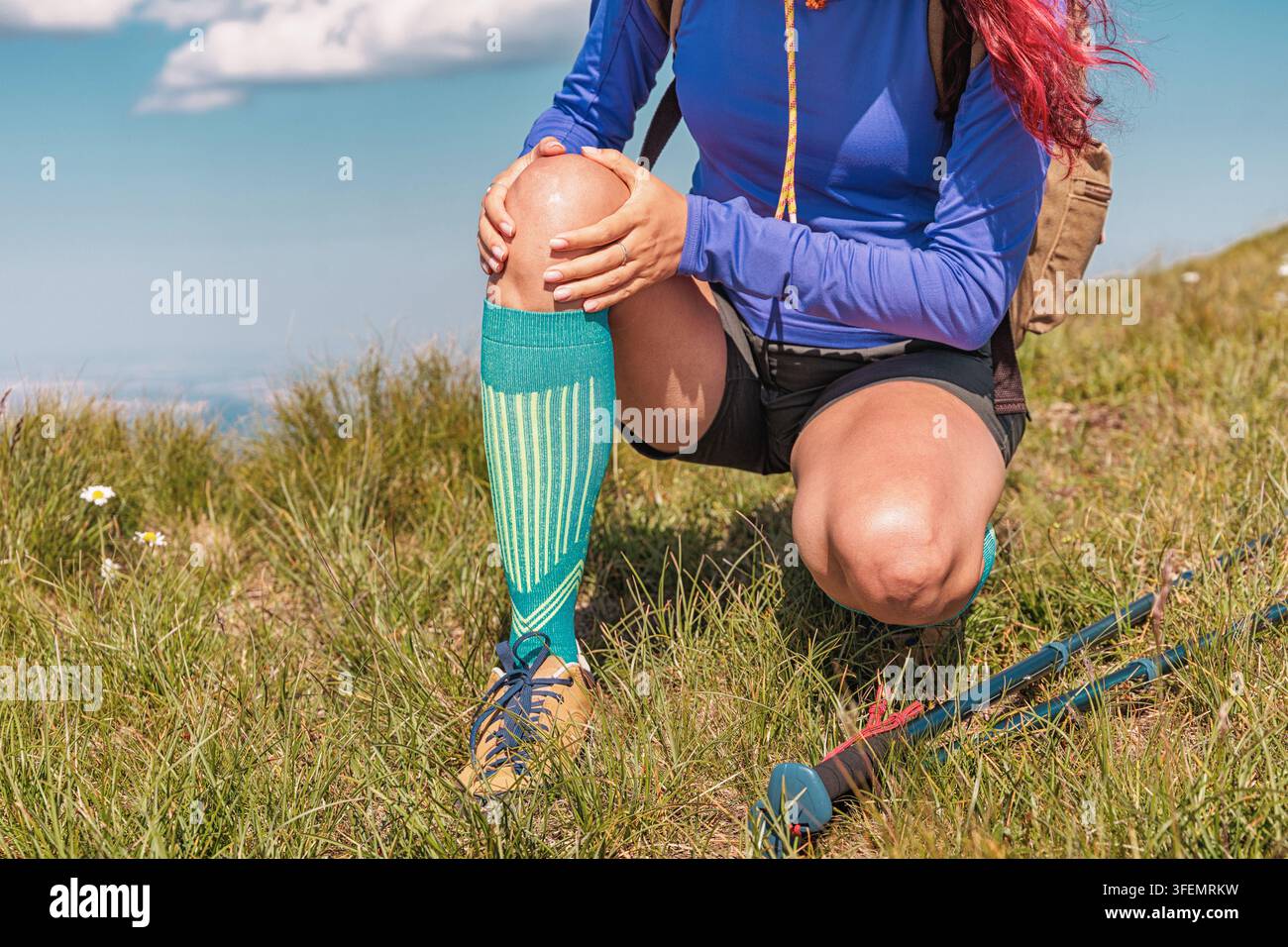 Escursionista che avverte dolori al ginocchio durante una pausa in una trekking in montagna, indossa calzini compressivi e attrezzatura da esterno Foto Stock