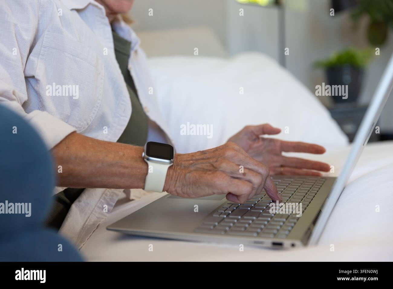 Donna anziana che digita su un laptop argentato mentre indossa uno smartwatch con cinturino bianco su lenzuola bianche Foto Stock