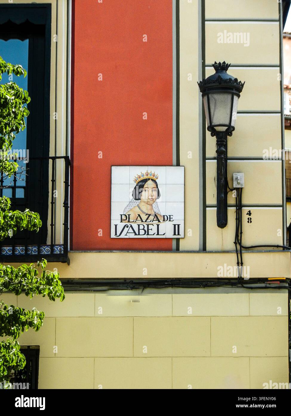 Segnale stradale „Plaza de Isabel II“ su una vivace parete arancione accanto a un lampione nero ornato nel centro storico di Madrid, Spagna. Foto Stock