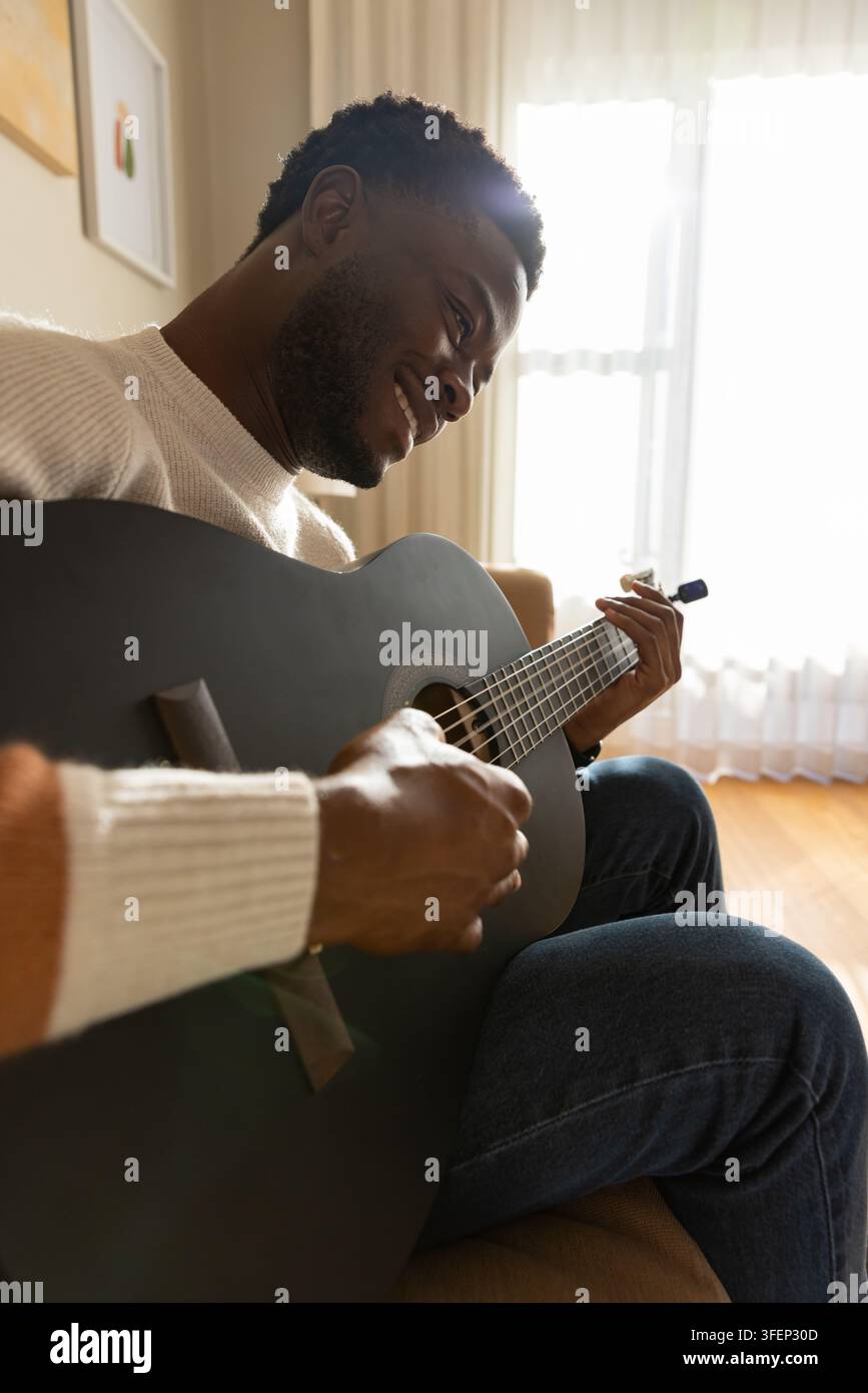 Uomo con maglione di lana color crema e jeans, che suona una chitarra acustica scura con capotasto sul divano Foto Stock