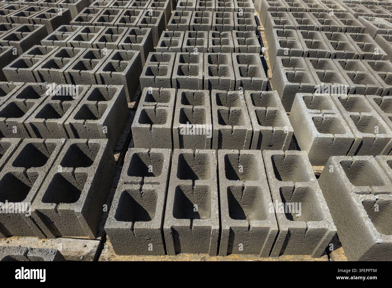Pile organizzate di blocchi cavi di cemento sono collocate sotto la luce solare in un cantiere edile. Le ombre forti sottolineano le superfici ruvide del bui Foto Stock
