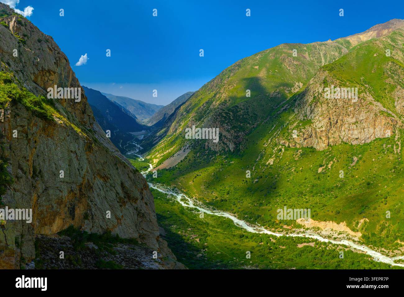 Inquadratura ampia che cattura una maestosa valle montana con scogliere rocciose e pendici verde vivace. Un fiume sereno scorre attraverso il paesaggio sotto un blu chiaro Foto Stock