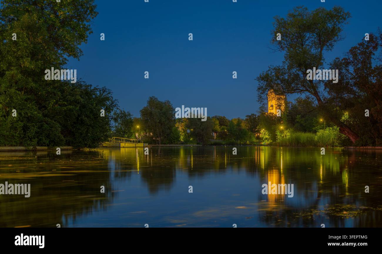 Il padiglione del parco Assiniboine risplende calorosamente al crepuscolo sopra la superficie immobile dello stagno delle anatre a Winnipeg, Manitoba. Una fitta chioma degli alberi incornicia l’acqua Foto Stock