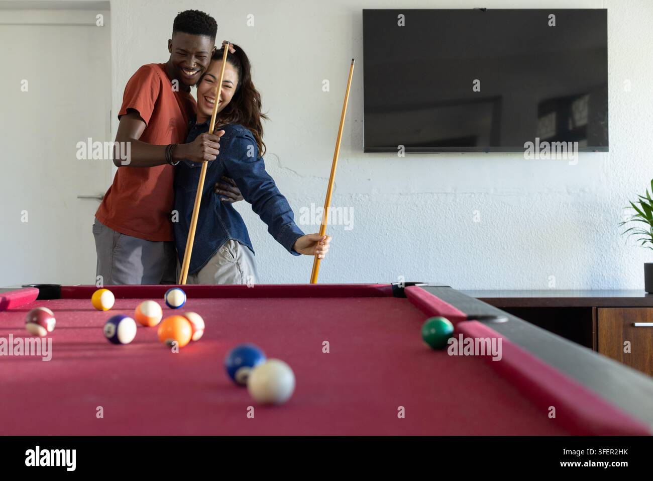 Coppia che sorride e si abbraccia tenendo le stecche da biliardo su un tavolo rivestito di feltro rosso con palle da biliardo a casa Foto Stock