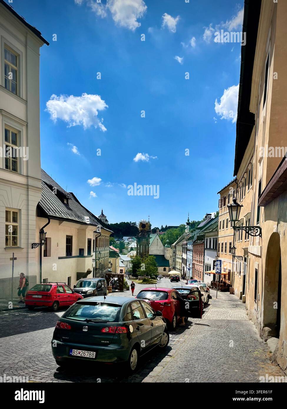 Foto verticale che guarda giù una strada acciottolata verso la Piazza Trinity a Banská Štiavnica, Slovacchia, con auto parcheggiate e case storiche - Immagine stock catturata con smartphone