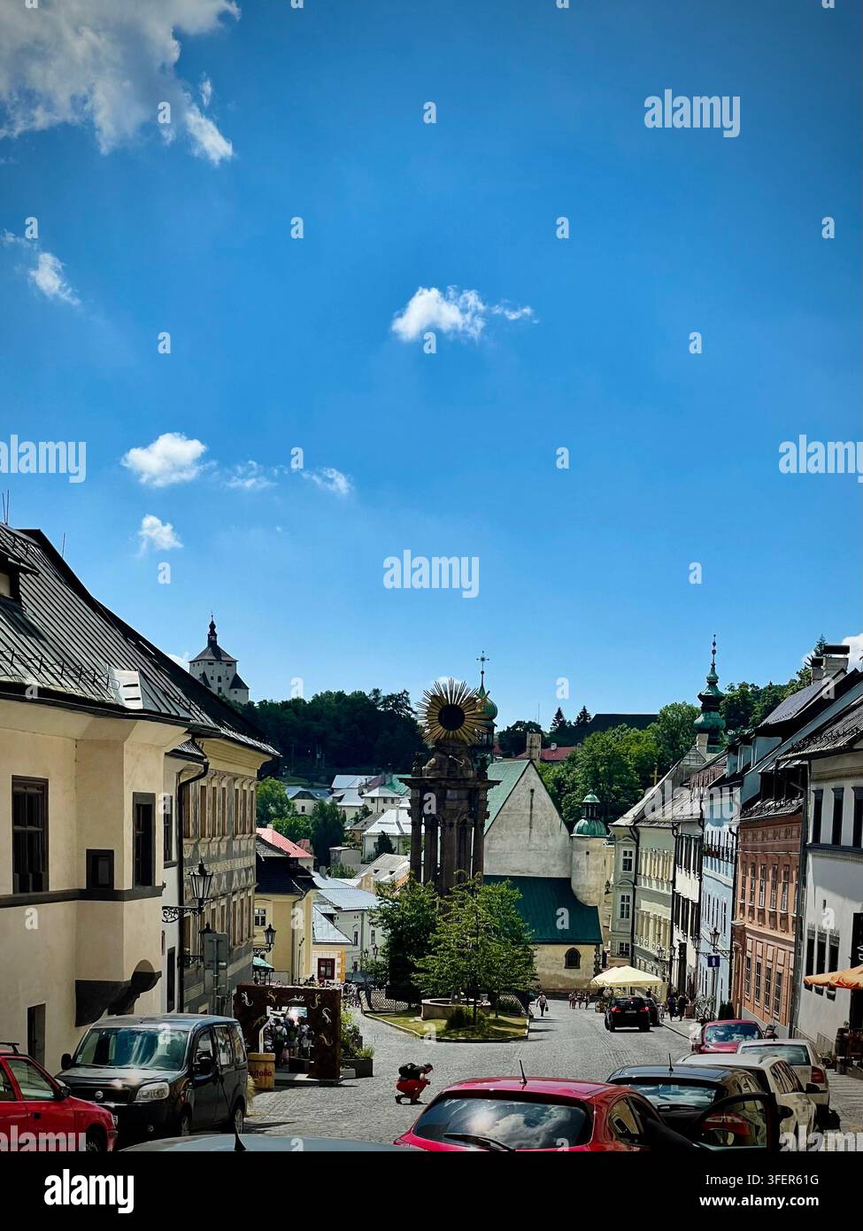 Foto verticale che guarda giù una strada di ciottoli verso Trinity Square e la Colonna della Santa Trinità a Banská Štiavnica, Slovacchia, incorniciata da elementi storici - Immagine stock catturata con smartphone
