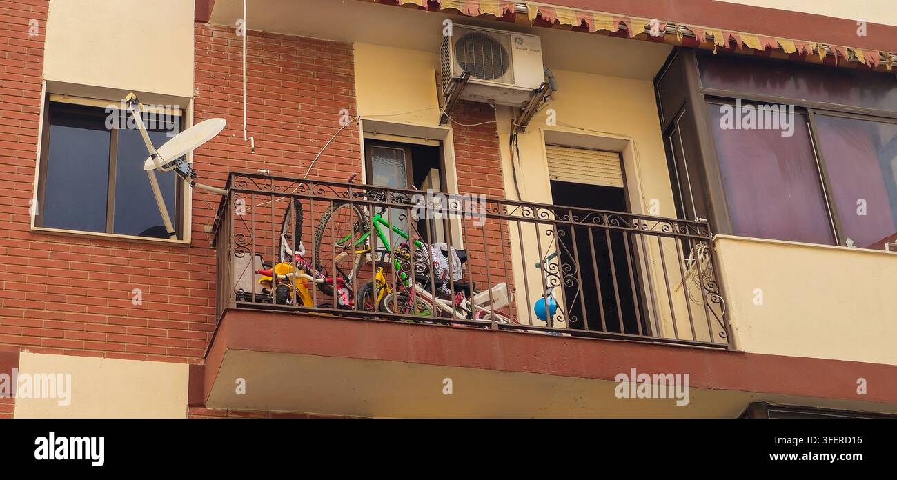 Balcone di appartamento quotidiano affollato di biciclette per bambini, giocattoli e oggetti domestici accanto a una parabola satellitare e un condizionatore a Baeza, Spagna. Foto Stock