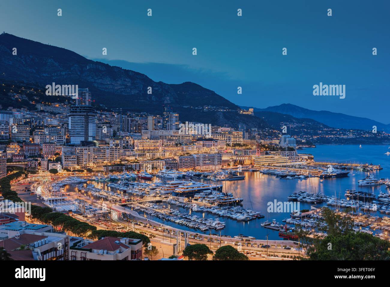 Vista aerea del Port Hercules a Monaco al crepuscolo, con yacht di lusso ormeggiati nel porto, edifici cittadini illuminati e montagne sullo sfondo Foto Stock