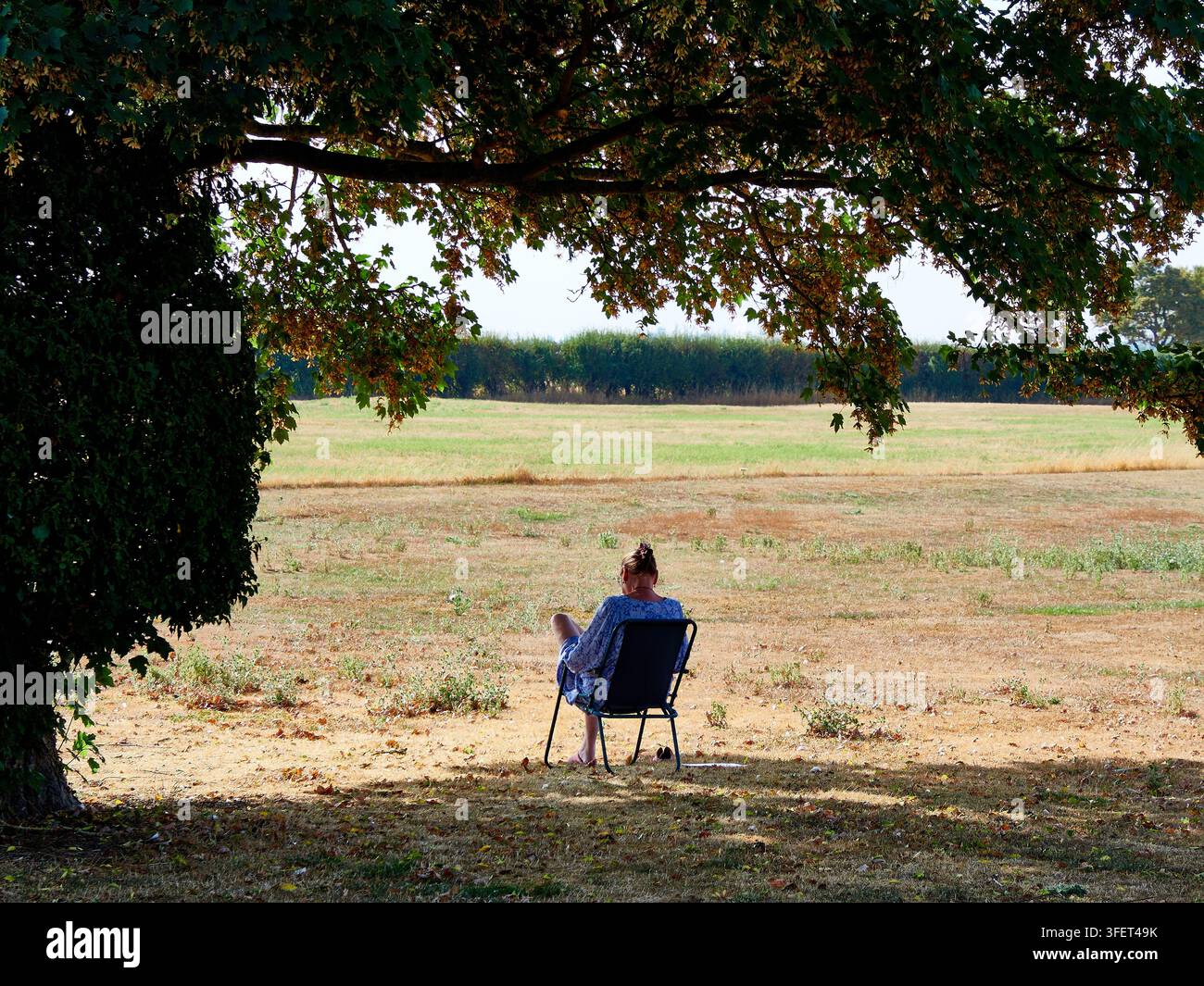 Donna seduta su una sedia all'ombra che si affaccia su un campo arido di campagna — solitudine estiva tranquilla Foto Stock