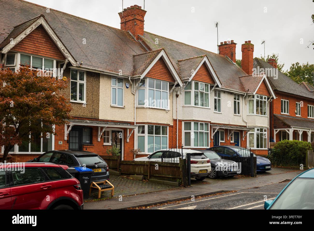 Una fila di case a schiera di periferia a Northampton con giardini anteriori, vialetti e diverse auto parcheggiate lungo una strada residenziale bagnata in una giornata nuvolosa Foto Stock