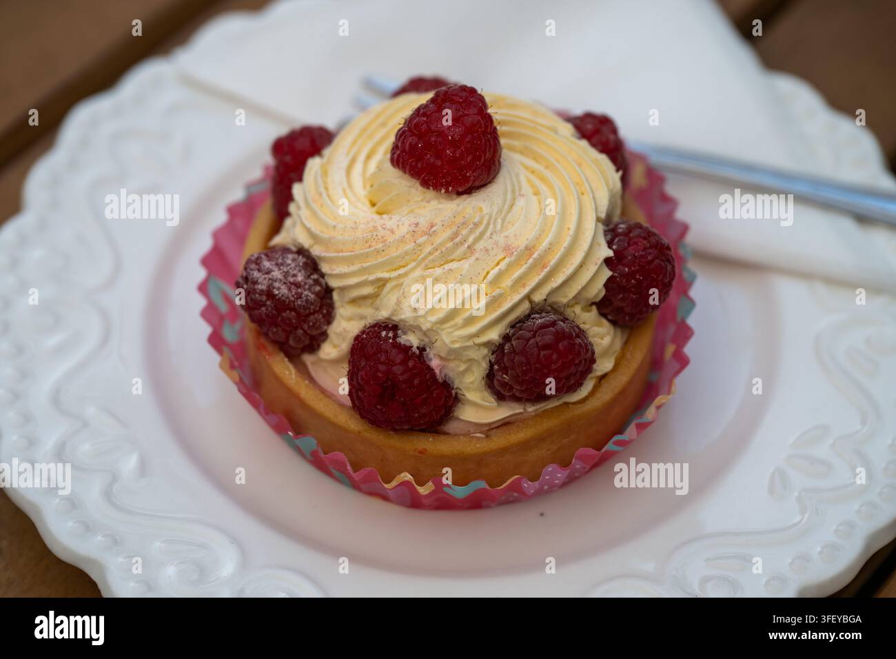 Una tartina alle frutta in miniatura ripiena di panna montata alla vaniglia e ricoperta di lamponi freschi è posata in una coppetta di carta rigata su un dolce bianco ornato Foto Stock