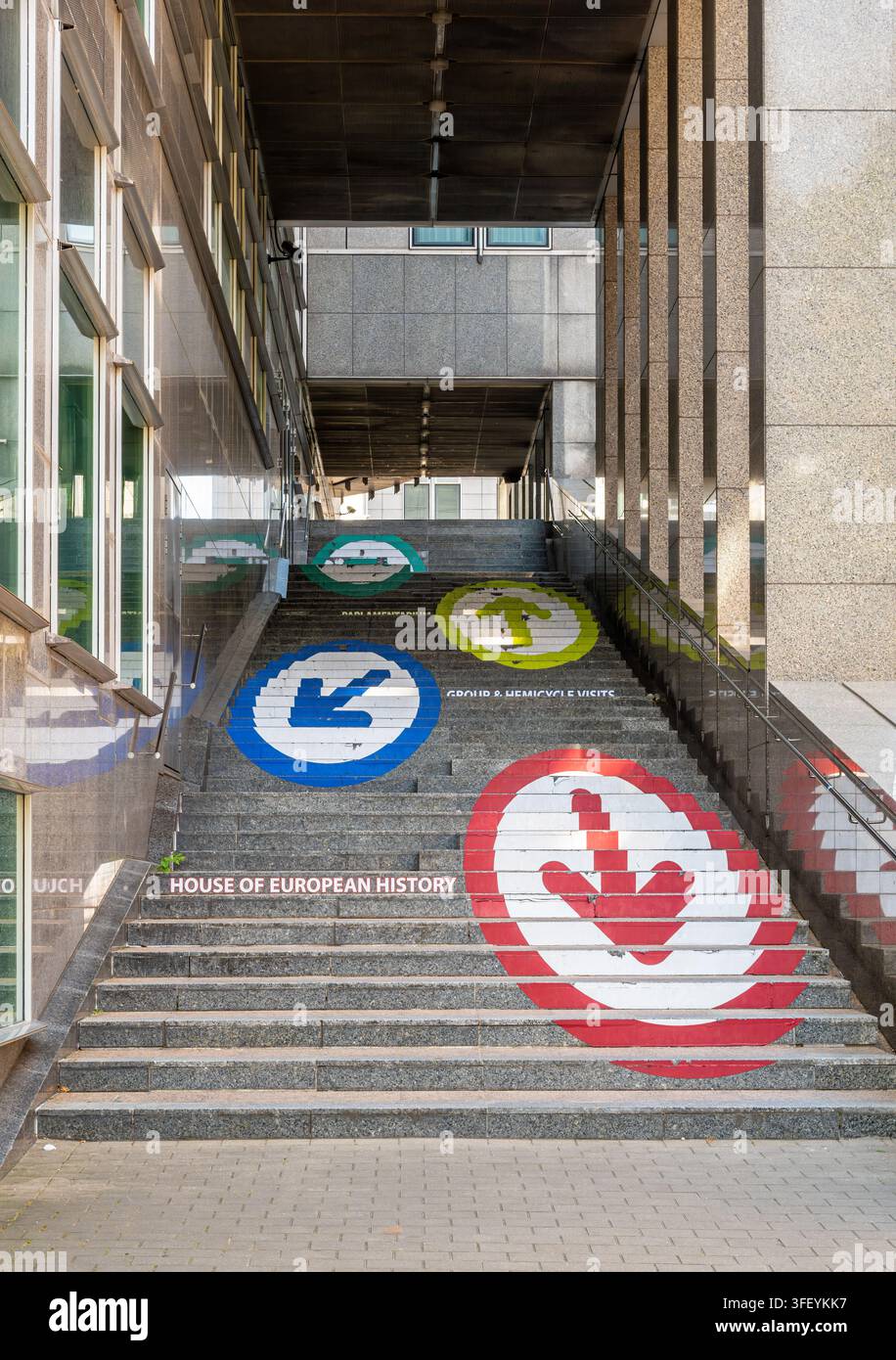 Bruxelles, Belgio – 8 agosto 2026: Ampia scala in cemento del Parlamento europeo dipinta con simboli direzionali colorati, incorniciata da un'alta colonna in pietra Foto Stock