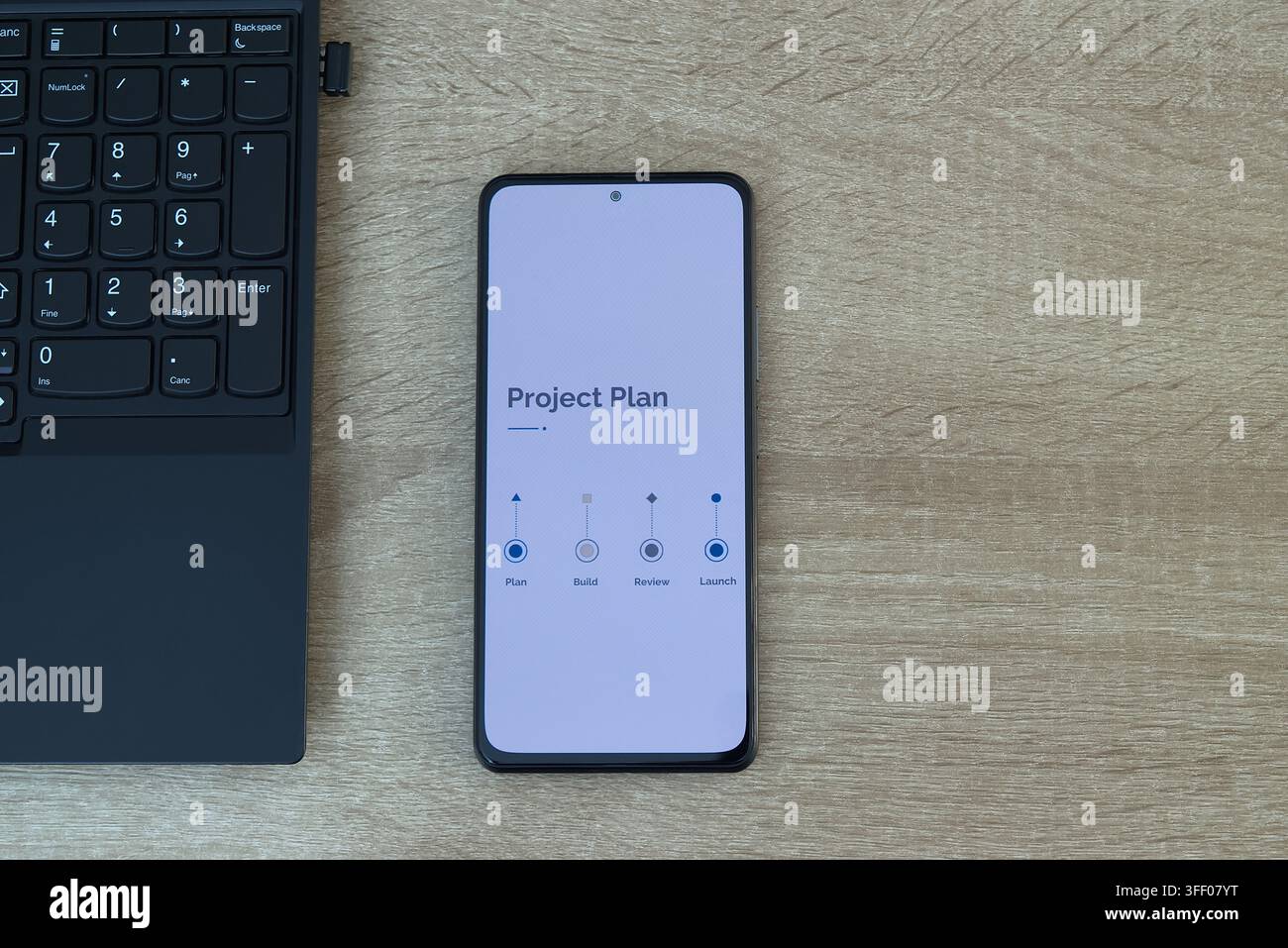 Vista dall'alto di uno smartphone che mostra un'interfaccia di piano di progetto pulita con fasi che includono piano, costruzione, revisione e lancio, posizionato accanto a una tastiera su Foto Stock