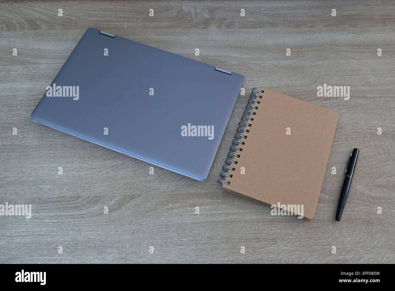 Vista angolare superiore minimalista di un computer portatile grigio chiuso posizionato accanto a un quaderno kraft a spirale e a una penna nera elegante su una superficie di legno neutra Foto Stock