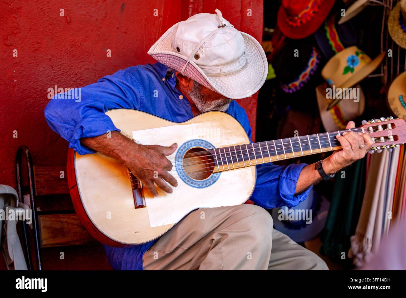 SAN MIGUEL DE ALLENDE, MESSICO  01 NOVEMBRE 2019: Un uomo suona la chitarra davanti a un muro rosso. Cappelli colorati pendono sullo sfondo. Foto Stock