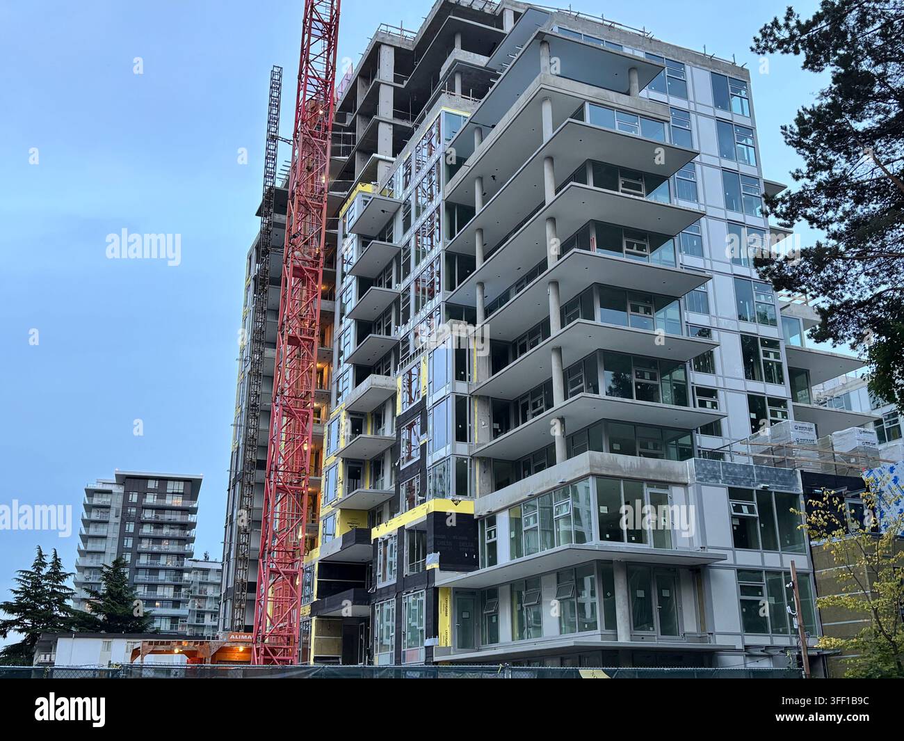 Un moderno palazzo residenziale di grandi dimensioni in costruzione, con una gru rossa imponente e balconi in vetro curvi ed eleganti contro un cielo nuvoloso - Immagine stock catturata con smartphone