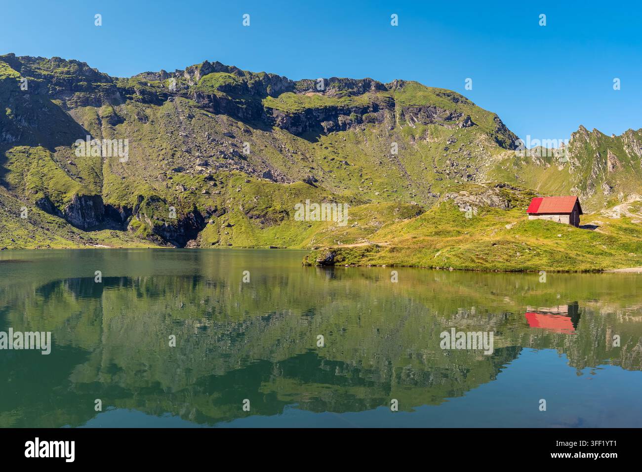 Piccola capanna rossa sul tetto accanto al lago Balea con riflessi delle montagne sotto un cielo blu limpido in Romania Foto Stock
