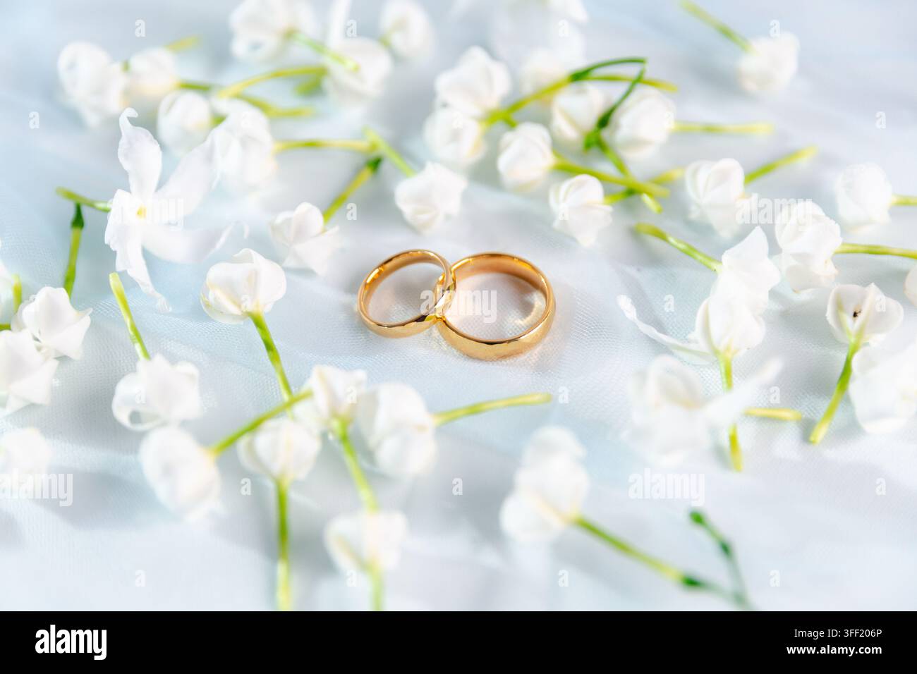 Due fedi nuziali d'oro circondate da delicate fiori bianchi su un morbido tessuto bianco, creando una scena nuziale elegante e romantica che simboleggia l'amore, il matrimonio Foto Stock