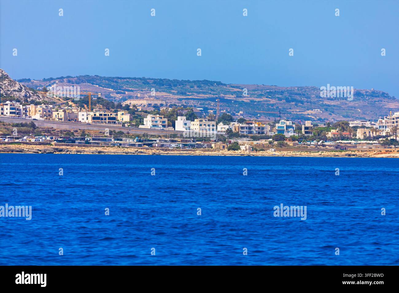 L'insediamento costiero di Bahar ic Caghaq a Malta si trova annidato lungo la costa rocciosa. Edifici moderni bianchi costellano il paesaggio, contro lo sfondo di un'arida zona ondulata Foto Stock