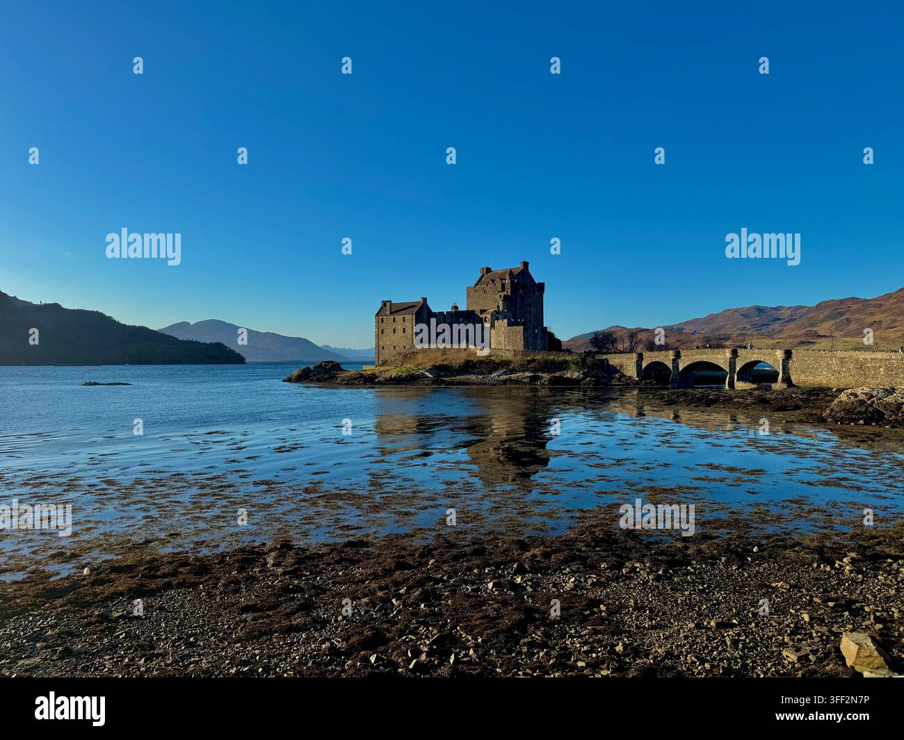 Castello di Eilean Donan, Highlands scozzesi, sotto un cielo blu limpido, con il suo ponte di pietra e le montagne circostanti che si riflettono nell'acqua calma. Foto Stock
