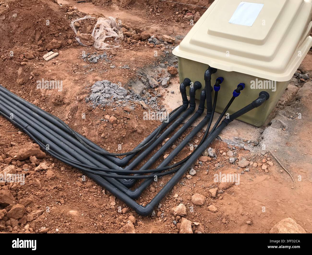 Sistema sotterraneo che mostra più tubi neri e cavi collegati a un'unità di distribuzione esterna, posizionata in terreno rosso scavato durante una casa Foto Stock