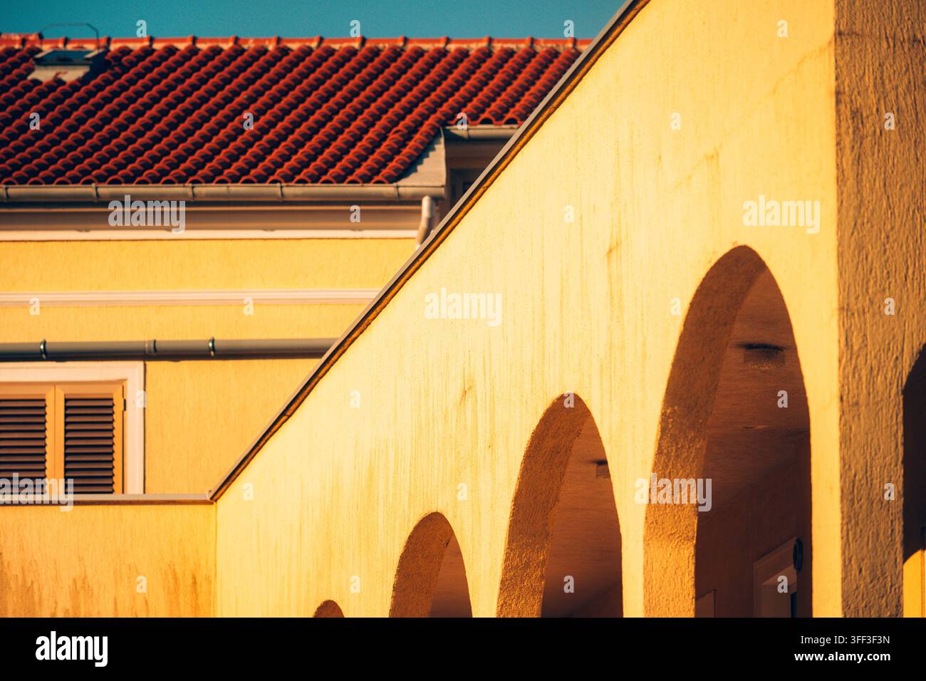 Archi architettonici gialli caldi con un tetto in tegole di terracotta rossa e una finestra con imposte illuminata dalla luce dorata. Messa a fuoco selettiva. Foto Stock