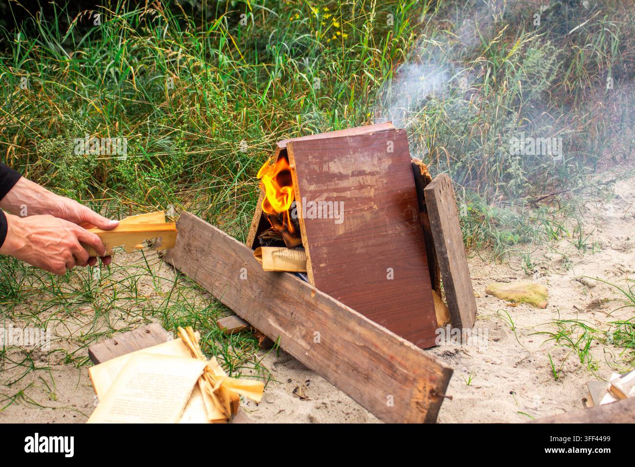 Giovane uomo in comodi abiti da trekking accende un falò all'aperto usando bastoni e vecchie assi di legno. Immagine perfetta per il campeggio, il turismo e il relax attivo Foto Stock