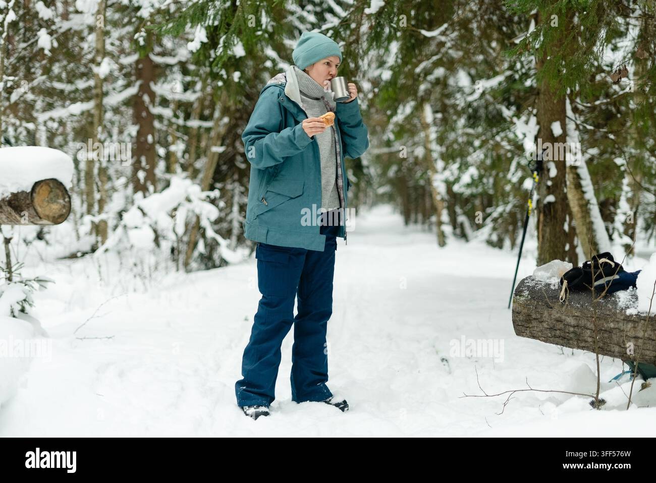 Donna sulla quarantina che fa una pausa invernale con una bevanda fumante e un dolce in una foresta innevata. Donna matura vicino a un ceppo con una tazza calda e una dolcezza. Pausa tranquilla all'aperto durante una fredda passeggiata invernale Foto Stock