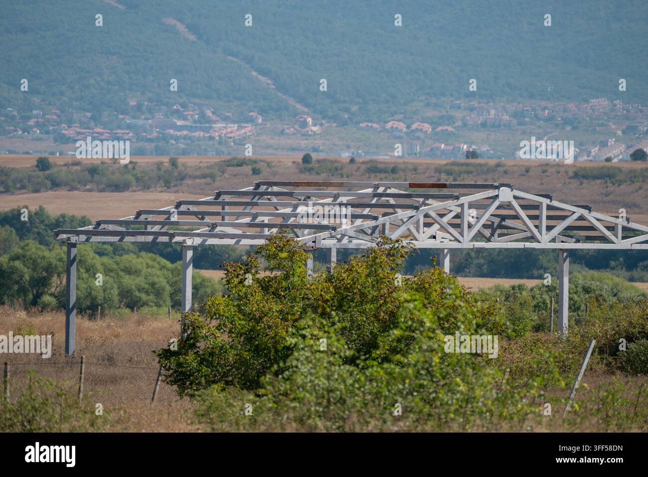 La struttura scheletrica bianca in acciaio di un edificio incompiuto si erge in un campo rurale secco e invaso dalla vegetazione, con una città e una grande montagna nella distanza nebbiosa Foto Stock