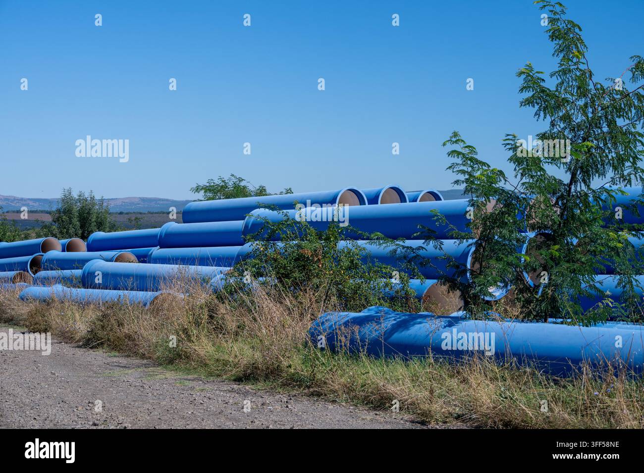 Grandi tubi industriali blu sono impilati lungo una strada sterrata in un campo di erba secca, con alcuni piccoli alberi verdi che crescono tra di loro, contro uno sfondo Foto Stock