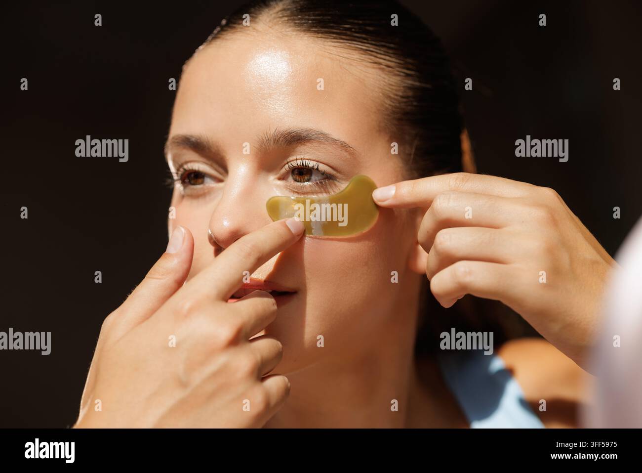 Giovane donna applica idrogel sotto il cerotto per gli occhi a casa, evidenziando la routine di cura del viso per l'idratazione, il sollievo dalle occhiaie e l'illuminazione delle occhiaie scure Foto Stock
