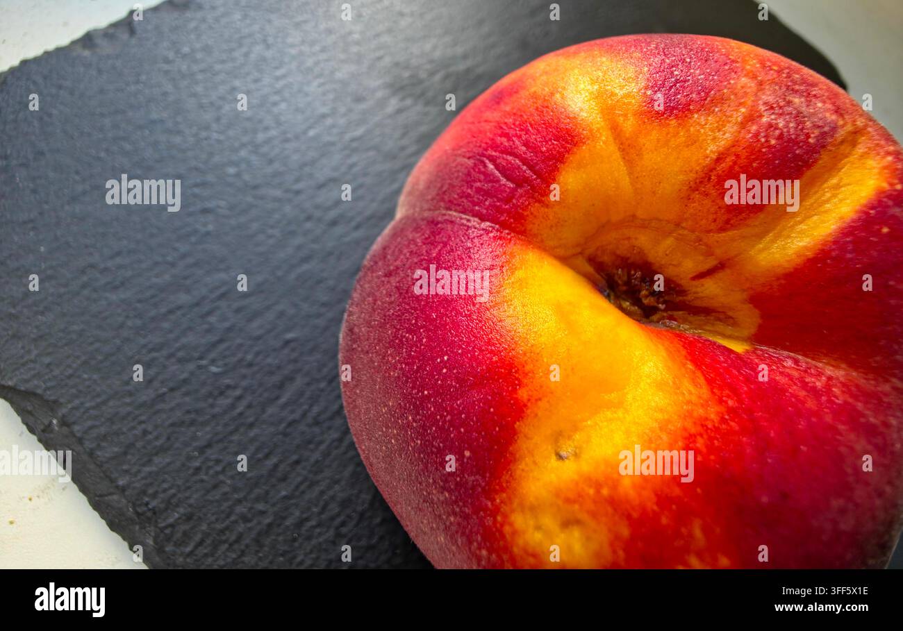 Un pesche maturo con tonalità calde di giallo e rosso si posa su una superficie di ardesia scura. La sua pelle liscia luccica nella luce soffusa mostrando la sua perfezione succosa e Foto Stock