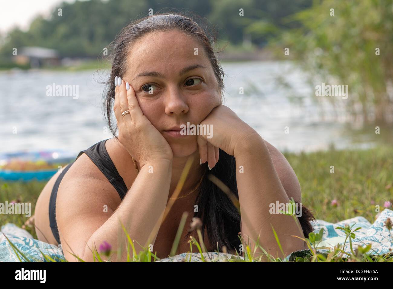 Una donna pensierosa appoggia il viso tra le mani mentre è sdraiata su una coperta vicino all'acqua. Foto Stock