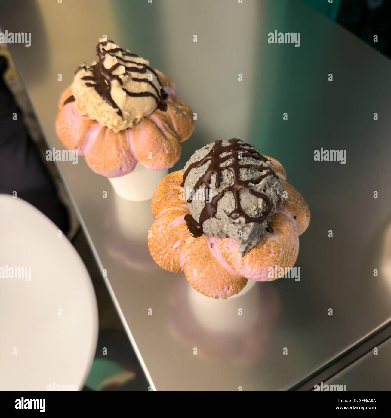 Due paste uniche a forma di fiore ripiene di gelato cremoso, generosamente ricoperte di sciroppo al cioccolato su un bancone metallico lucido. Foto Stock