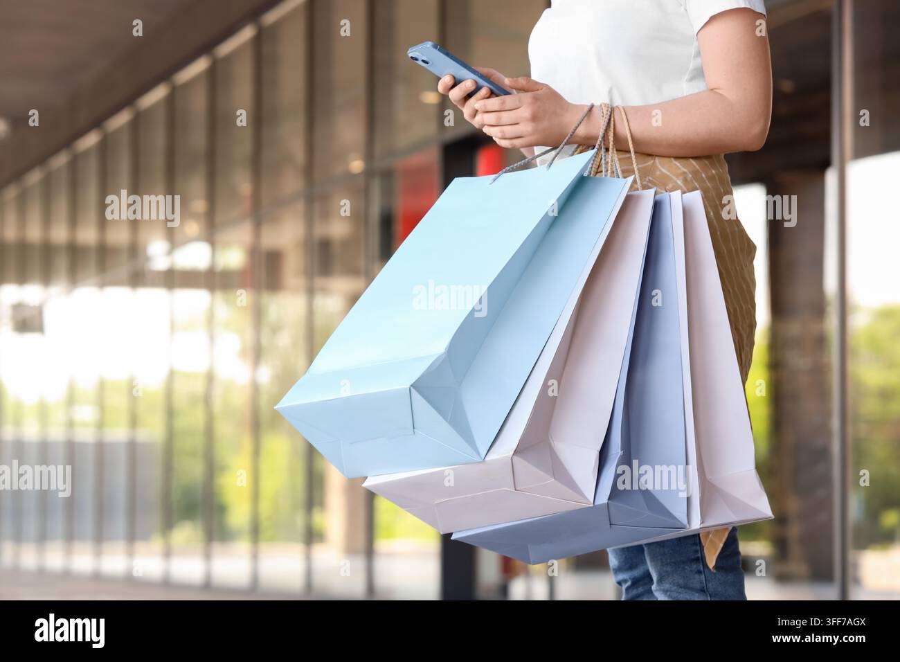 Comportamento dei consumatori e acquisti al dettaglio, donna che utilizza uno smartphone mentre porta borse di carta fuori da un centro commerciale moderno per vendite stagionali e tendenze della moda Foto Stock