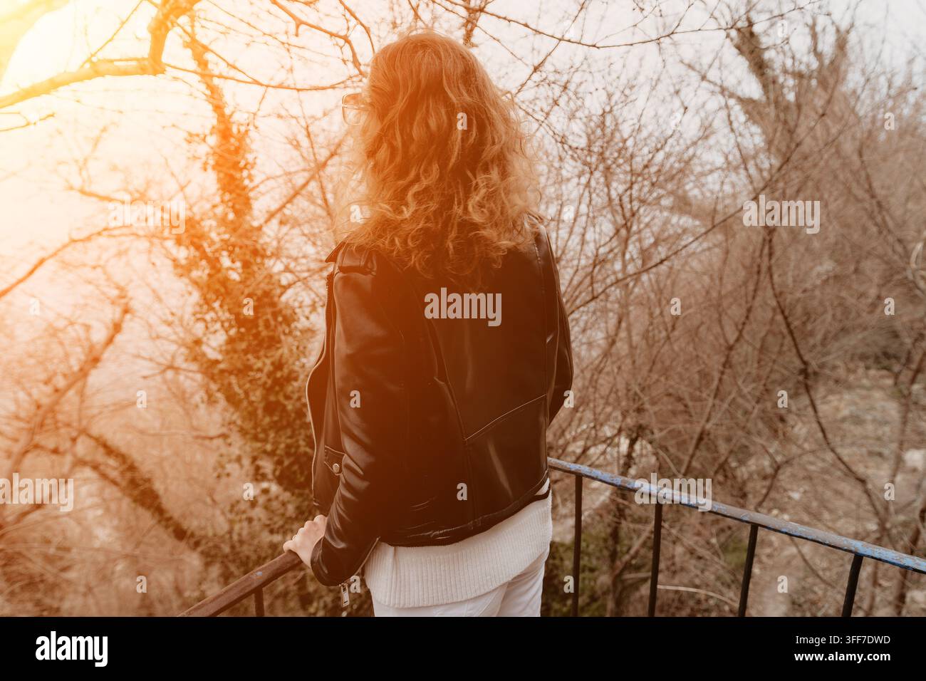 Donna su un balcone nella natura, donna con una giacca di pelle che guarda una foresta spoglia da un balcone rustico al tramonto Foto Stock