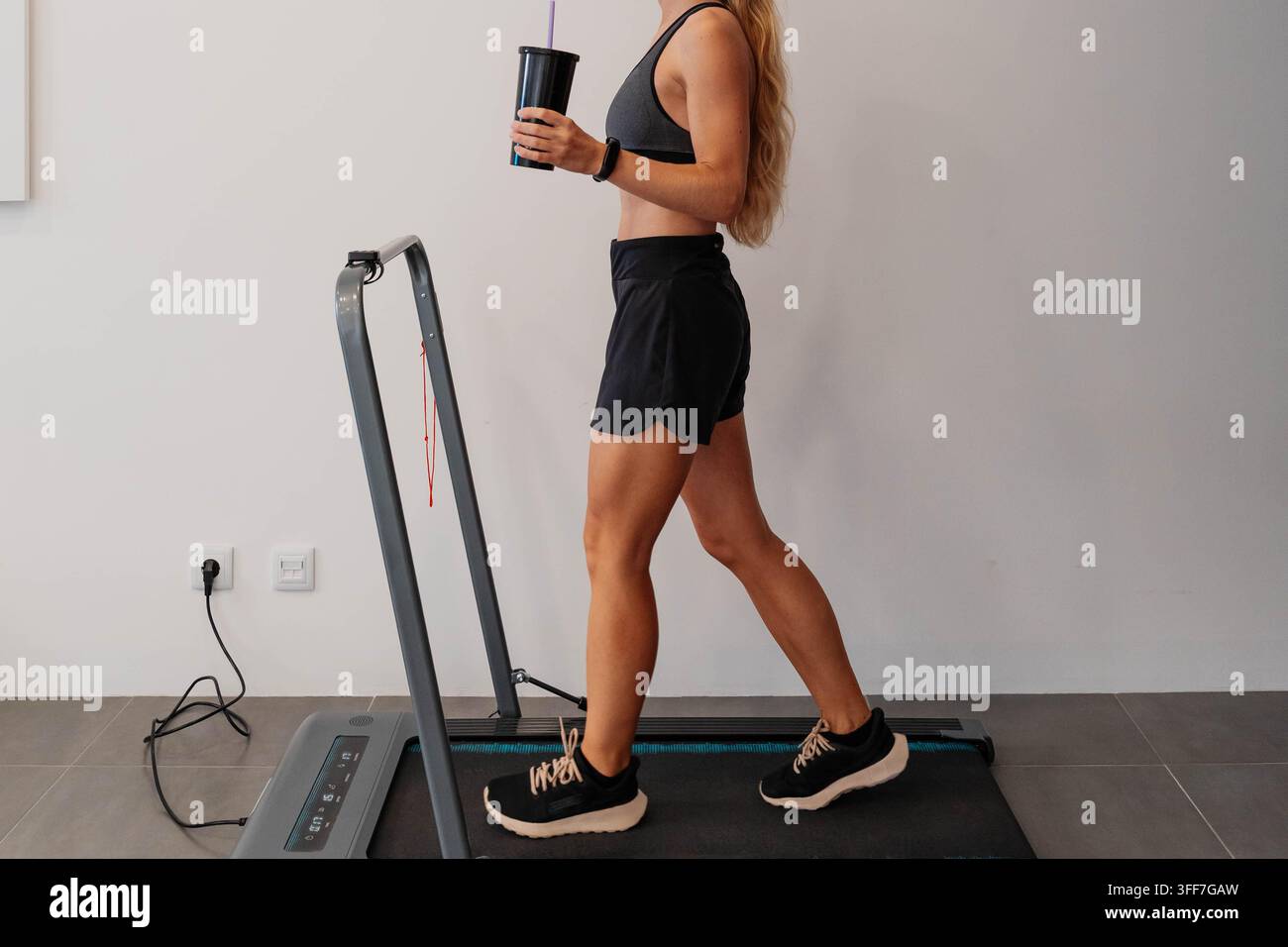Donna in abbigliamento sportivo cammina su un tapis roulant pieghevole compatto a casa mentre tiene un tumbler con una cannuccia e indossa un fitness tracker. Cardio a basso impatto Foto Stock