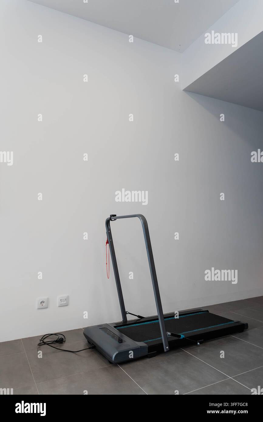 Tapis roulante pieghevole compatto con corrimano si trova accanto a una presa di corrente in una stanza vuota con pareti bianche e pavimento piastrellato. Attrezzatura per il fitness domestico per uso interno Foto Stock