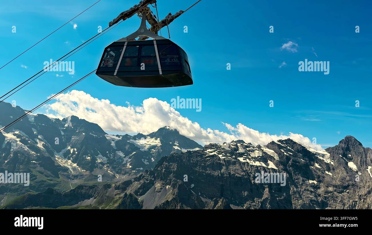 Funivia sopra aspre montagne alpine innevate. La funivia scivola sopra aspre montagne alpine con chiazze di neve sotto un cielo blu vivido e luminoso Foto Stock