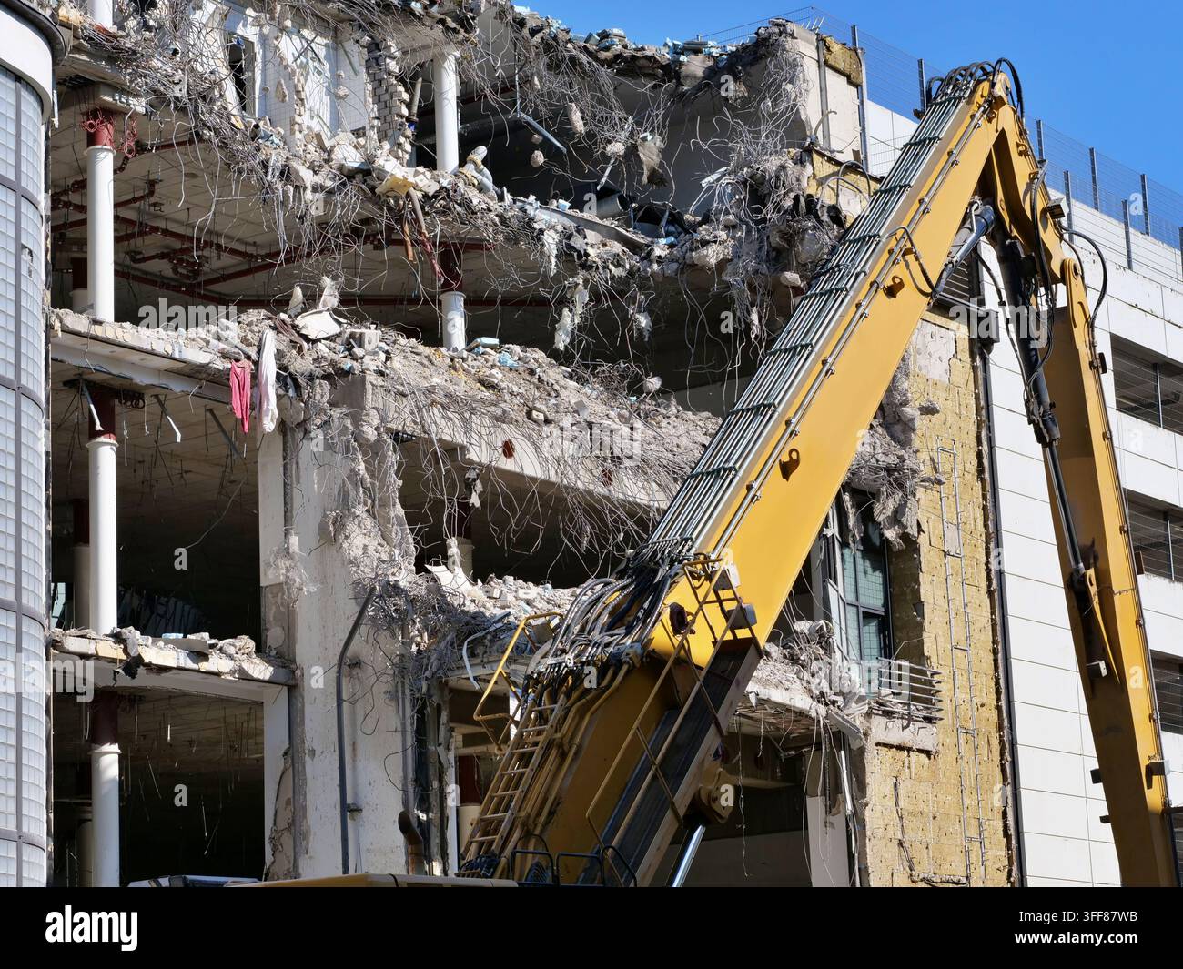 Un potente braccio di escavatore giallo ad alta portata posizionato davanti a un edificio a più piani durante una demolizione strutturale importante. Lastre di cemento esposte, t Foto Stock